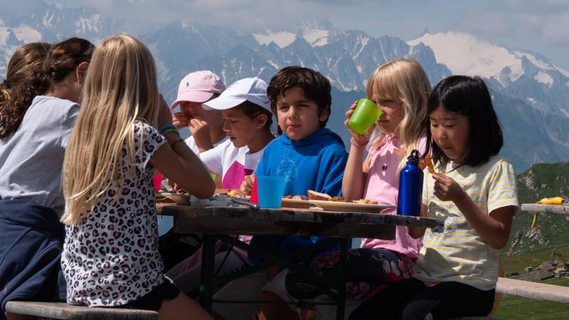 Group of international young kids having lunch outside during the 2024 summer camp of Les Elfes in Verbier, Europe